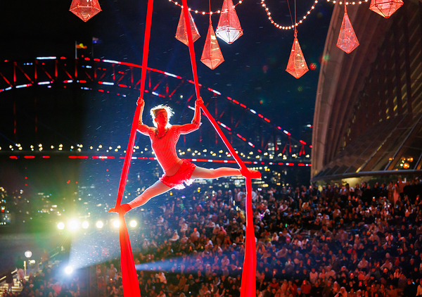 An aerial performer in a red costume executes a split on red silks during a Vivid Sydney 2026 event, with the Sydney Harbour Bridge and Opera House illuminated in the background and a large audience watching An aerial performer in a red costume executes a split on red silks during a Vivid Sydney 2026 event, with the Sydney Harbour Bridge and Opera House illuminated in the background and a large audience watching